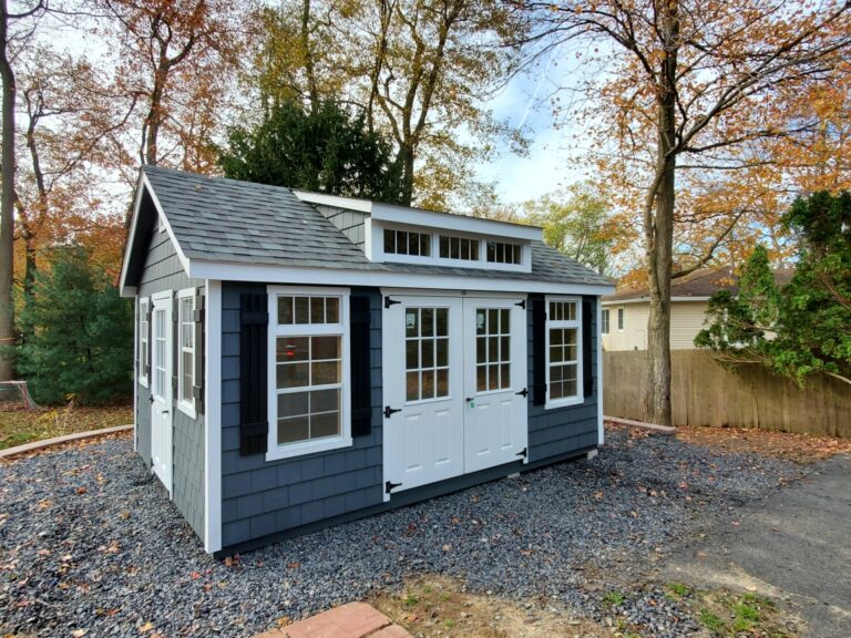 Blue-gray shed with white trim and a dormer, standing on a crushed stone pad in a wooded backyard