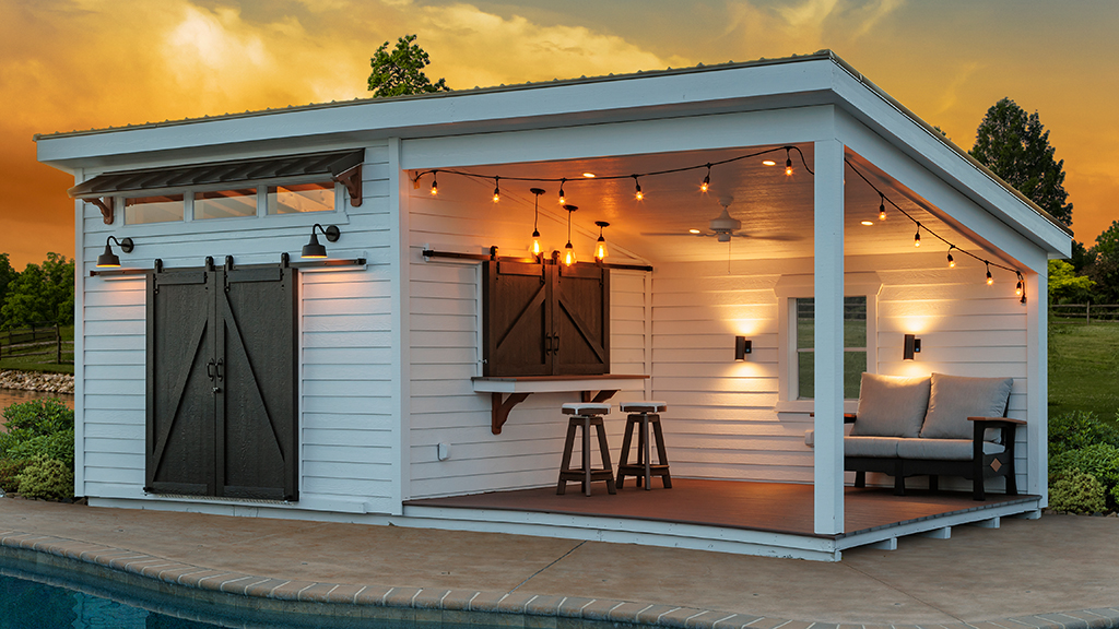 White pool house at dusk with sliding barn doors, a pass-through serving bar and a covered lounge area beside a pool