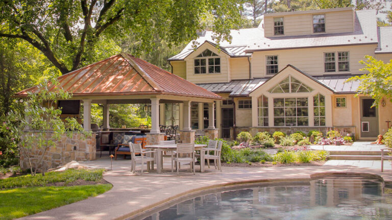 Hip-roof pavilion with a copper metal roof, stone fireplace and outdoor kitchen beside a backyard pool